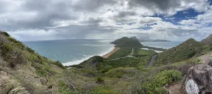 Panoramic view from Timothy Hill over the St. Kitts Southeast Peninsula, with ocean on both sides, winding roads, green hills and Nevis beneath clouds in the distance.