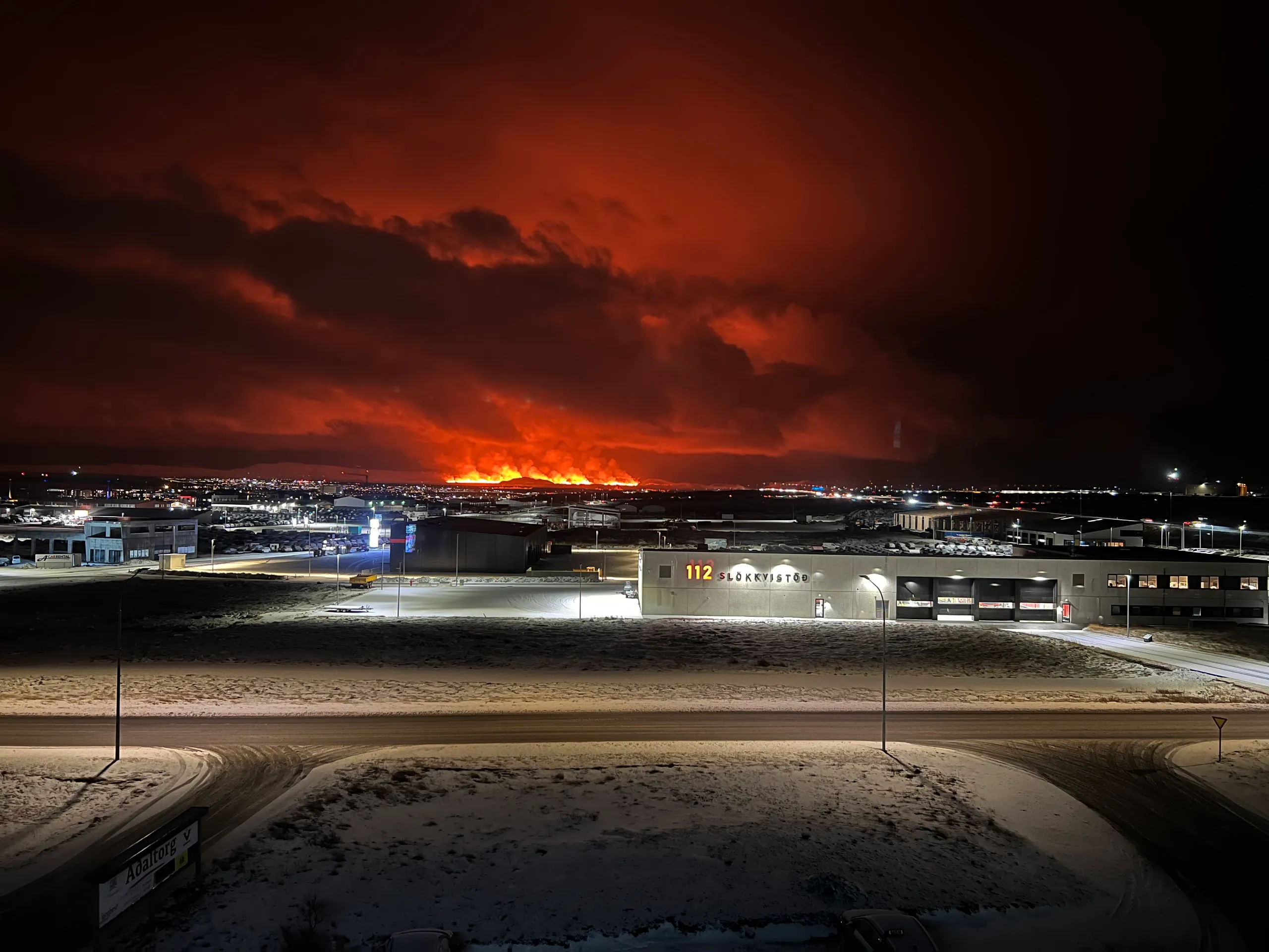 Orange lava and an illuminated volcanic plume on the horizon beyond buildings near Keflavík at night.