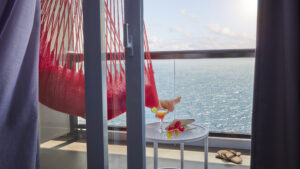 A sailor relaxes in a red hammock on Scarlet Lady’s balcony overlooking the sea, with a cocktail on the table.