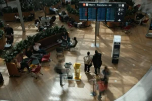 Travelers wait beneath an airport departures board, with luggage and terminal seating nearby.