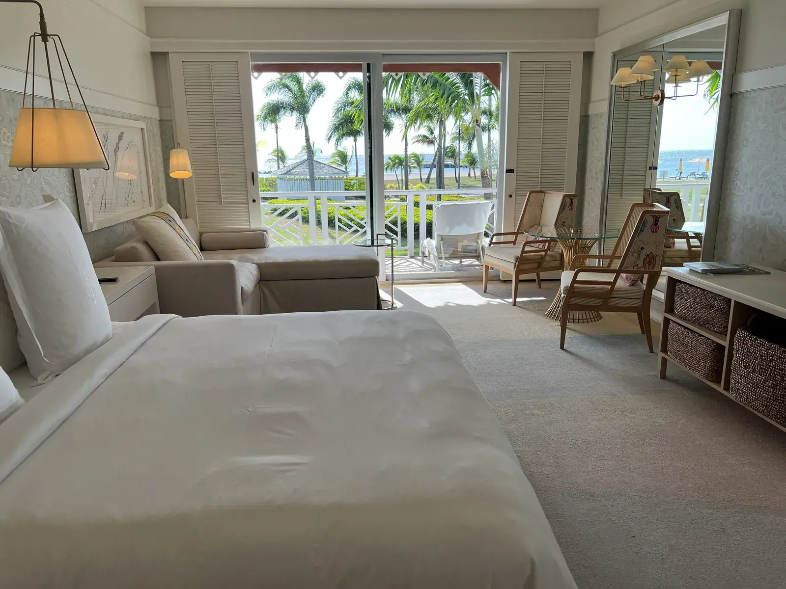 Bed, sofa and chairs beside balcony doors with palms and sea outside.