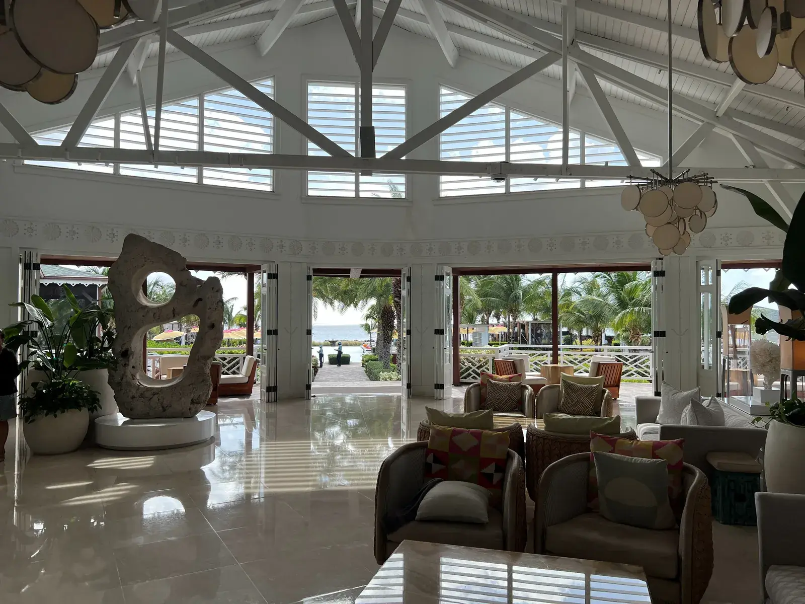 White roof beams, lounge seating and open doors framing palms and sea.