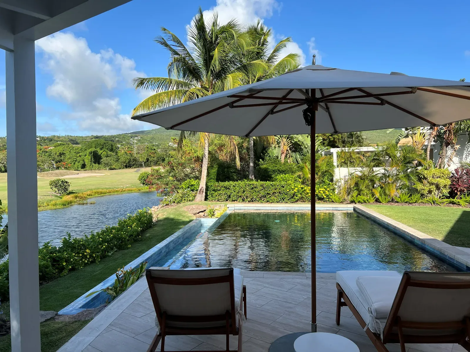 Umbrella and two loungers facing a small pool with a pond and green grounds beyond.