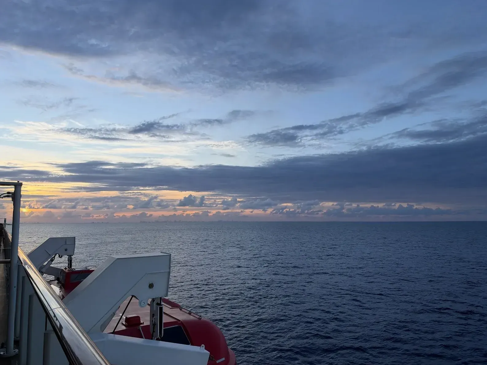 Sunset over the sea from Norwegian Getaway, with the ship’s rail and lifeboat in view.